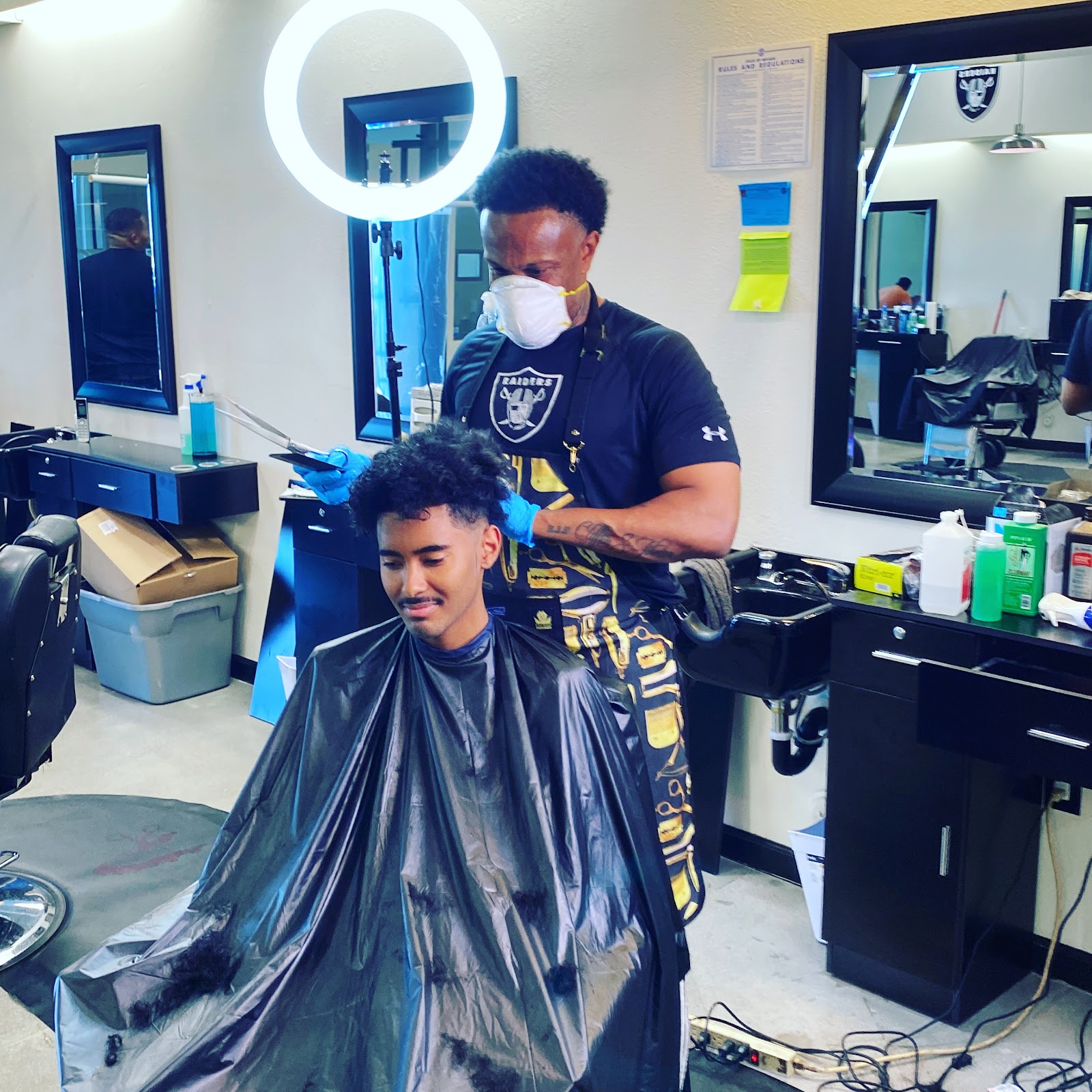 Masked barber cutting a client's curly hair under a ring light