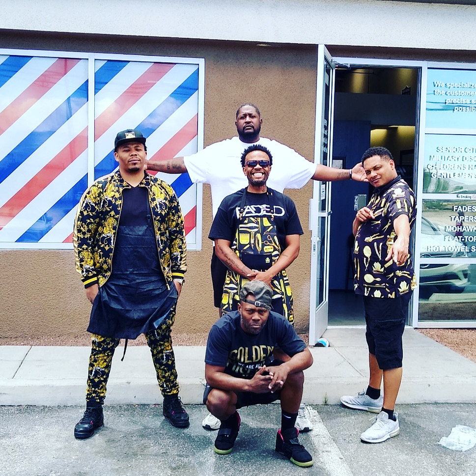 Five barbers posing together outside the Faded Barbershop storefront with barber pole windows